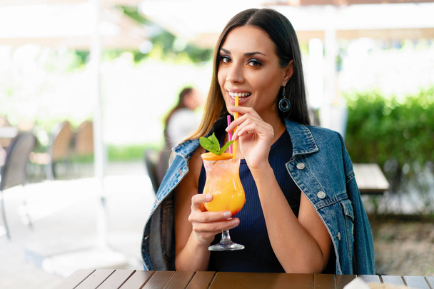 ein schönes junges Mädchen von europäischem Aussehen trinkt einen exotischen Sommercocktail, während sie an einem Tisch eines Straßencafés sitzt. bekleidet mit Jeansjacke - Foto, Bild