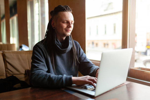 Young Businessman with dreadlock having doing his work in cafe near the big window with laptop. Youth culture. Neformal guy. Remote works. IT specialist - Fotografie, Obrázek