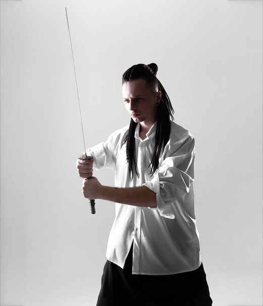 Young man dressed in the white shirt with dreadlocks holding a samurai sword. Glamour photo. Studio shooting. business man with Japanes sword - Foto, Imagem