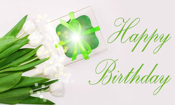 Happy Birthday μήνυμα σημάδι στο φως φόντο. Λευκά λουλούδια τουλίπας και κουτί δώρου με πράσινη κορδέλα πλώρη επίπεδη θέσει. Λουλούδι μπουκέτο συγχαρητήρια ευχετήρια κάρτα. Αντιγραφή προβολής banner ιστοσελίδας χώρου. - Φωτογραφία, εικόνα