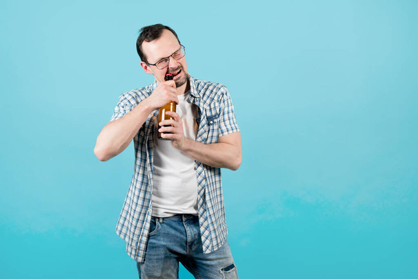 a man in a plaid shirt with bristles in glasses diligently opens a bottle of beer with his teeth - Foto, Imagen
