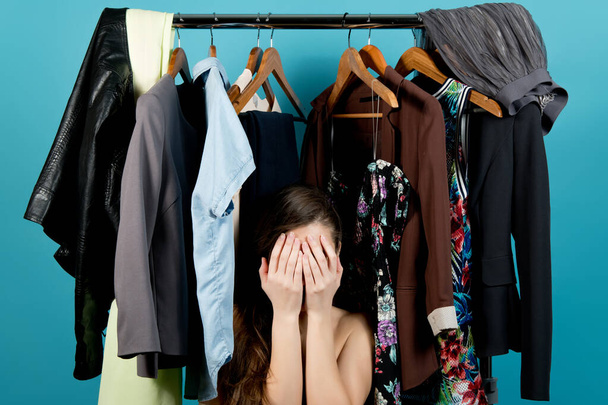 Girl covers her face, lost in the choice of clothes on the background of hangers with clothes, blue background - Φωτογραφία, εικόνα