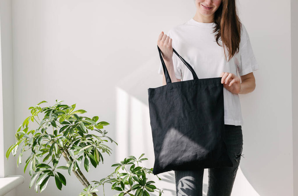 Young woman holding black textile eco bag against white wall. Ecology or environment protection concept. White eco bag for mock up. - Fotoğraf, Görsel