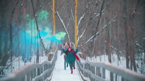 Two young happy women running on the snowy bridge holding smoke bombs - Záběry, video
