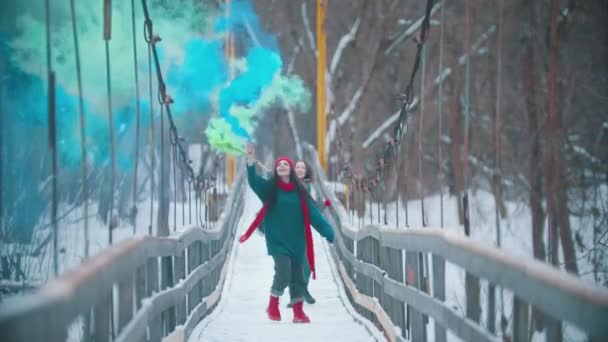 Két fiatal boldog nő szórakozik a havas hídon, színes füstbombákkal a kezükben. - Felvétel, videó