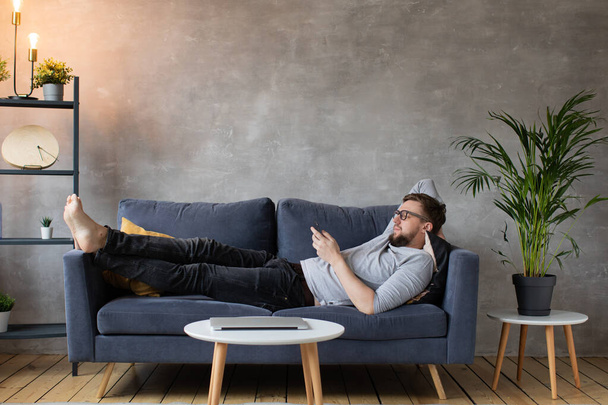 Jeune homme avec des lunettes est allongé sur le canapé et SMS au téléphone
. - Photo, image