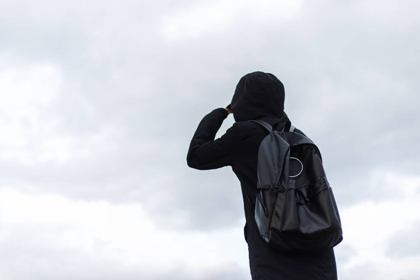 Vista posteriore del viaggiatore sullo sfondo del cielo, indossando cappotto nero e zaino
. - Foto, immagini