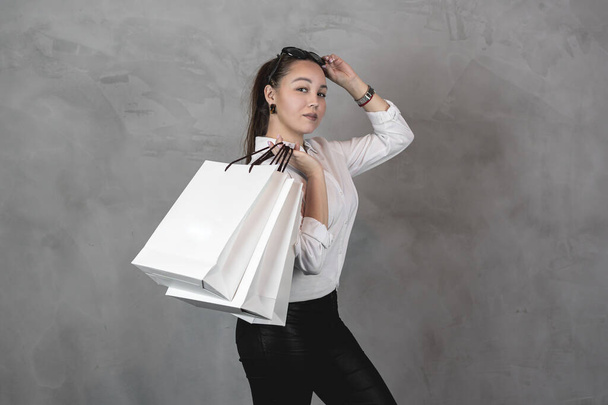 красива молода жінка в білій сорочці і сонцезахисних окулярах тримає сумки для покупок над сірим тлом справляється з простором
 - Фото, зображення