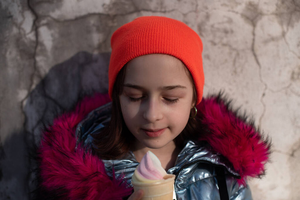 Leuk vrolijk peuter meisje in de winter hoed eten ijs. Meisje dat 's winters ijs eet. Portret van een meisje van 9 jaar oud. Schoolmeisje die buiten ijs eet op een warme zonnige winterdag. 10 jaar - Foto, afbeelding