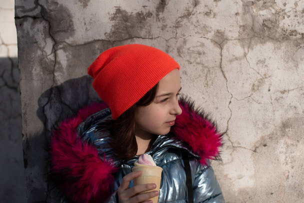 Cute Happy Maluch Dziewczyna w czapce zimowej jedzenia lodów. Dziewczyna jedząca lody zimą. Portret 9-letniej dziewczynki. Uczennica je lody na świeżym powietrzu w ciepły słoneczny zimowy dzień. 10 lat - Zdjęcie, obraz