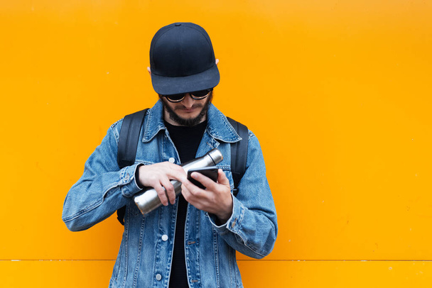 Porträt eines jungen selbstbewussten Hipster-Mannes auf orangefarbenem Hintergrund. Smartphone mit Thermo-Ökoflasche aus Stahl für Wasser in der anderen Hand. - Foto, Bild