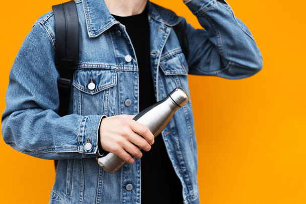 Male hand holding eco steel thermo bottle for water, wearing jeans jacket and backpack, on yellow background. - Fotoğraf, Görsel