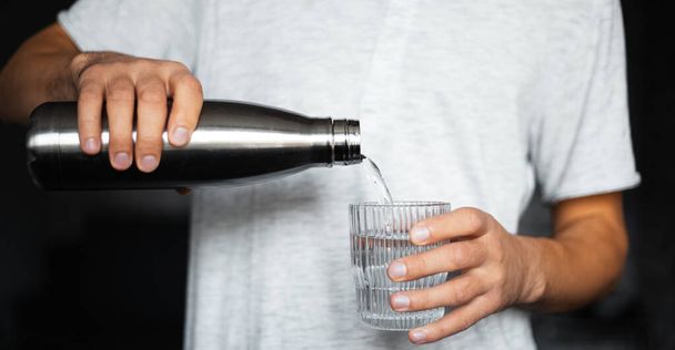 Male hands pours water into a glass from steel thermo eco bottle. - Фото, изображение