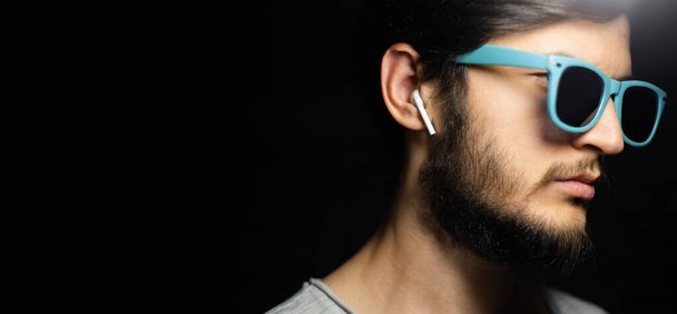 Portrait of young serious man with wireless earphones, looking up, wearing blue sunglasses, isolated on black background. - Fotografie, Obrázek