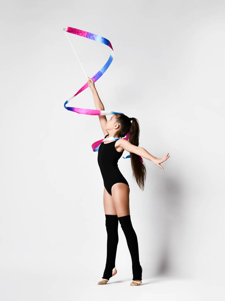Young girl gymnast in black sport body and uppers standing and making exercise with gymnastic tape over white background - Foto, immagini