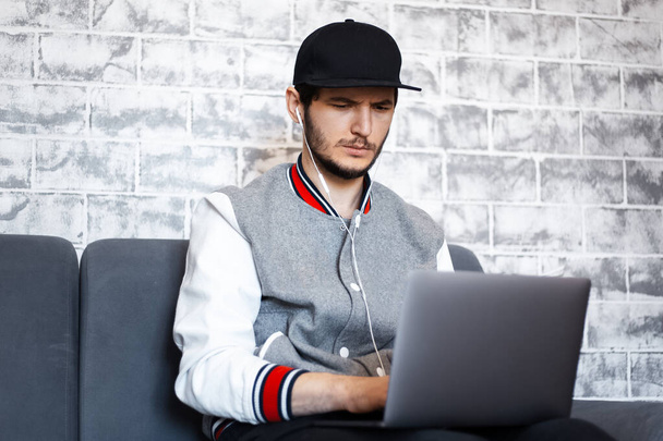Portret van een jonge freelancer, zittend op de bank met een kop koffie in de hand en laptop op benen. Grijze bakstenen muur achtergrond. - Foto, afbeelding
