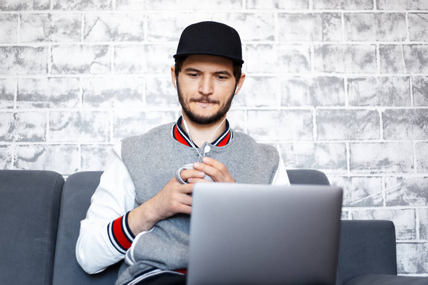 Serbest çalışan genç bir adamın portresi, elinde bir fincan kahve, bacaklarında dizüstü bilgisayarla kanepede oturuyor. Gri tuğla duvar arkaplanı. - Fotoğraf, Görsel