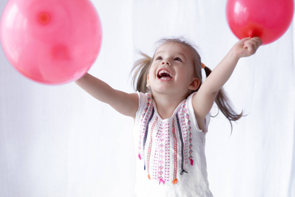 Happy and cheerful emotions of a little girl two years old. Little girl holds two red balls in her hands. Beautiful girl playing with balloons - Фото, зображення