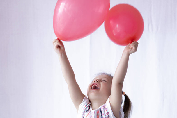 Ευτυχισμένα και χαρούμενα συναισθήματα ενός μικρού κοριτσιού δύο ετών. Το κοριτσάκι κρατάει δύο κόκκινες μπάλες στα χέρια της. Όμορφο κορίτσι παίζει με μπαλόνια - Φωτογραφία, εικόνα
