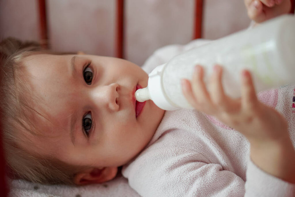 Klein mooi meisje gekleed in een pyjama ligt in een wieg. Een baby in een roze pyjama met een fles melk ligt in een wieg - Foto, afbeelding