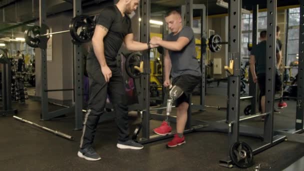 Eğitmen, spor salonunda protez bacakla egzersiz yapan genç adama yardım ediyor. - Video, Çekim