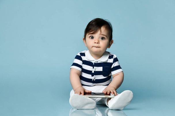 Toddler in striped t-shirt, white pants and booties. He holding smartphone, sitting on floor against blue background. Close up - Фото, изображение