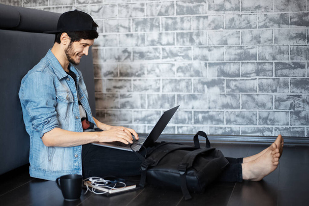 Retrato de jovem de jaqueta jeans e boné preto sentado no chão, trabalhando no laptop, usando smartphone, xícara preta de café
. - Foto, Imagem