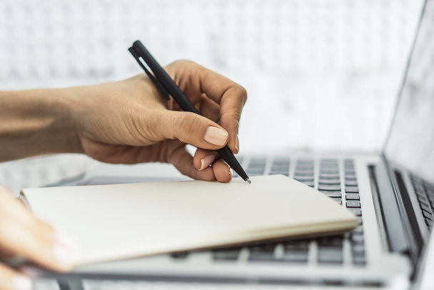 Mujer escribe con un bolígrafo en el diario en el teclado del ordenador portátil en una oficina soleada, negocio y concepto de educación. De cerca.
 - Foto, imagen