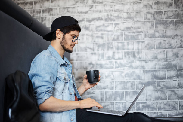 Портрет молодого чоловіка в джинсовій куртці і чорній кепці, що сидить на підлозі, працює на ноутбуці, використовуючи смартфон, чорну чашку кави
. - Фото, зображення