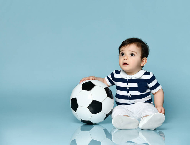 Niño en camiseta a rayas, pantalones blancos y botines. Está mirando hacia arriba, sentado en el suelo sobre fondo azul. De cerca.
 - Foto, imagen