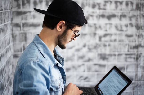 Portrait of young man looking in laptop with sunglasses and black cap on the background of grey brick wall. - Fotoğraf, Görsel