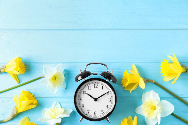 Black alarm clock with spring flowers and space for text on light blue wooden background, flat lay. Time change - Photo, image