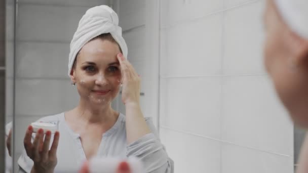 Adult woman with a towel on her head applying cream and looking in mirror enjoy natural beauty and healthy - Footage, Video