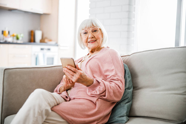 Smiling old lady sitting on couch and holding smartphone - Foto, Imagem