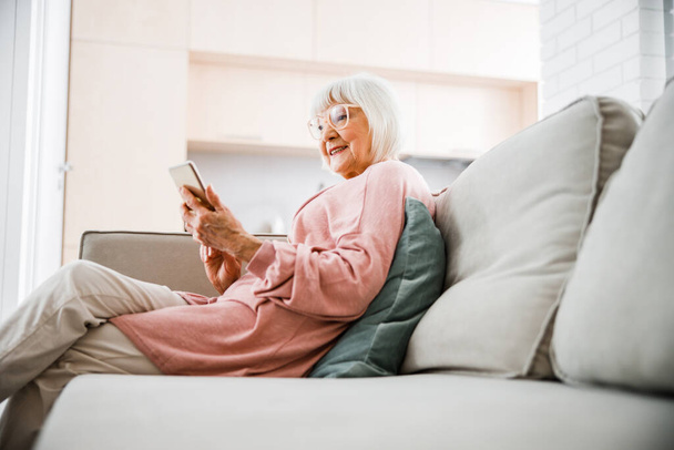 Joyful senior lady sitting on couch and using mobile phone - Foto, Imagen