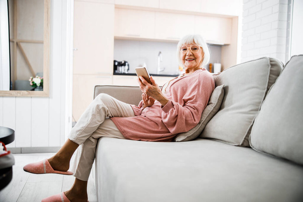 Smiling elderly woman sitting on couch and holding smartphone - Foto, Imagen