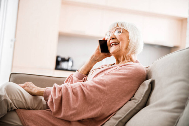 Vrolijke oude vrouw aan het praten op mobiele telefoon thuis - Foto, afbeelding