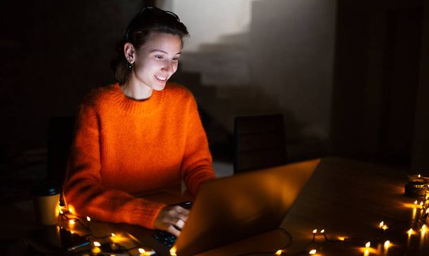 Portrait of smiling designer girl, working at graphic tablet on laptop. Wearing eyeglasses and orange sweater. In dark room home, with garlands. - Φωτογραφία, εικόνα