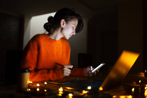 Retrato de la chica de diseño sonriente, trabajando en la tableta gráfica en el ordenador portátil. Usando anteojos y suéter naranja. En casa habitación oscura, con guirnaldas
. - Foto, Imagen