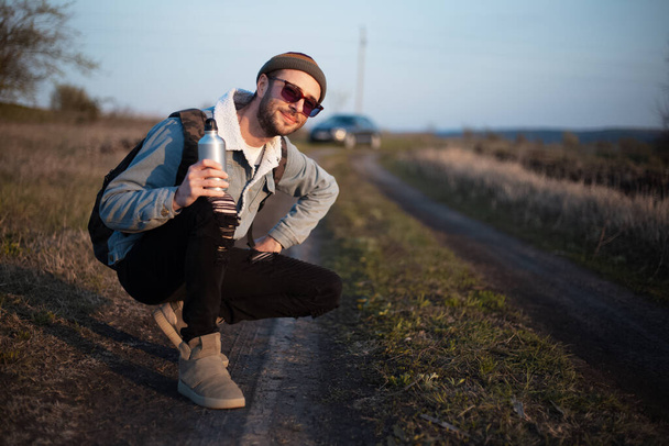 Portrait of young hipster man with backpack, smartphone and reusable aluminum water bottle outdoor. - Фото, изображение