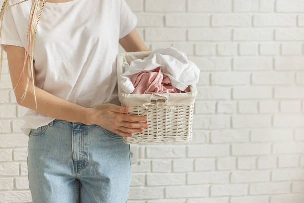 Woman holding wicker basket with heap of different clothes, on bricks wall background - Photo, Image