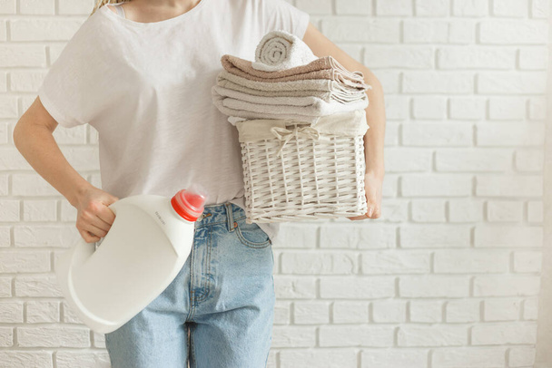 Жінка тримає пляшку гелю-миючого засобу та кошик з купою різного одягу перед пранням, на фоні стіни цегли
 - Фото, зображення