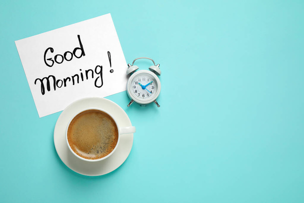 Delicious coffee, alarm clock and card with GOOD MORNING wish on light blue background, flat lay. Space for text - Foto, Imagem
