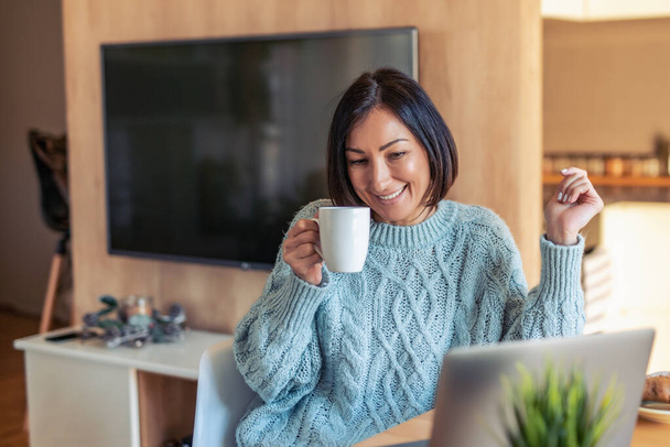 Hymyilevä nainen työskentelee kannettavan tietokoneen parissa ja juo kahvia kotitoimistossa
. - Valokuva, kuva