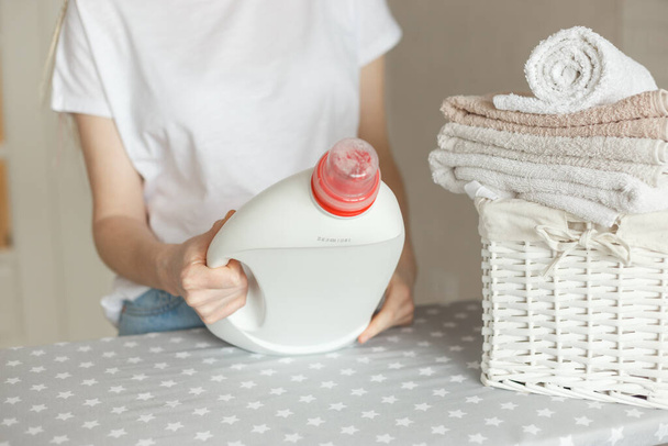 Frau hält weiße Flasche mit Waschmittel in der Hand. Weidenkorb mit Kleidung und auf dem Tisch - Foto, Bild