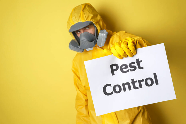 Man wearing protective suit with insecticide sprayer holding sign PEST CONTROL on yellow background - Foto, Bild