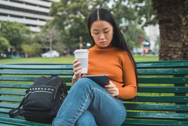 Portrait of young Asian woman using her digital tablet while holding a cup of coffee in the park outdoors. - Photo, image