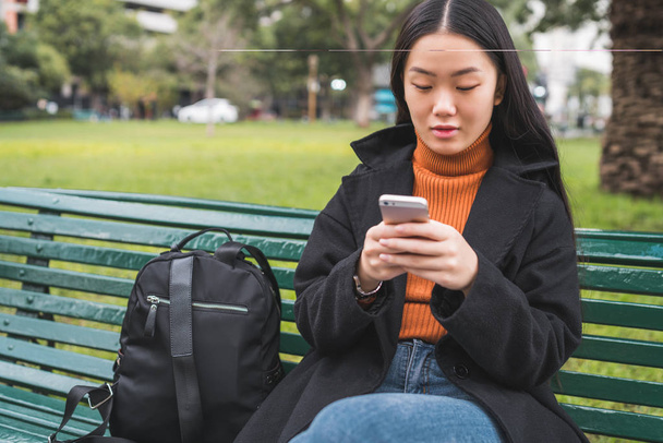 Portrait of young Asian woman using her mobile phone while sitting on a bench in the park. Communication concept. - Valokuva, kuva