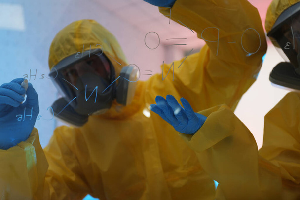Scientists in chemical protective suits writing formula on glass board at laboratory. Virus research - Photo, Image