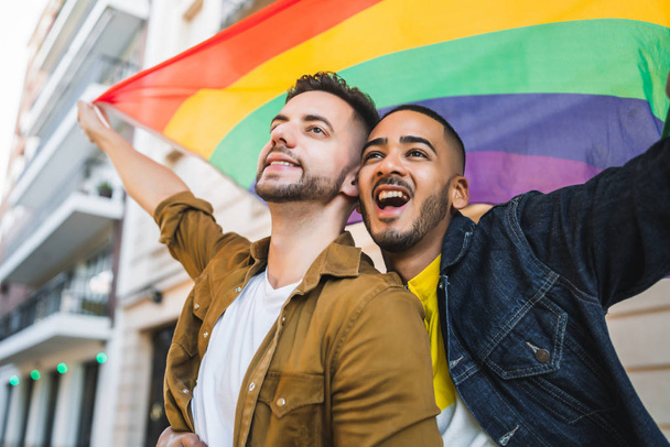 Portrait of young gay couple embracing and showing their love with rainbow flag at the street. LGBT and love concept - Φωτογραφία, εικόνα